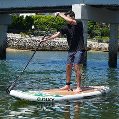 Person paddleboarding on a Nixy board in a body of water with a bridge and greenery in the background.