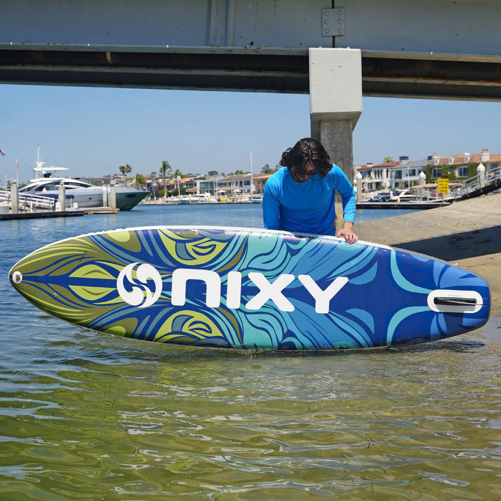 Person holding a NIXY surfboard by the water with a bridge and buildings in the background