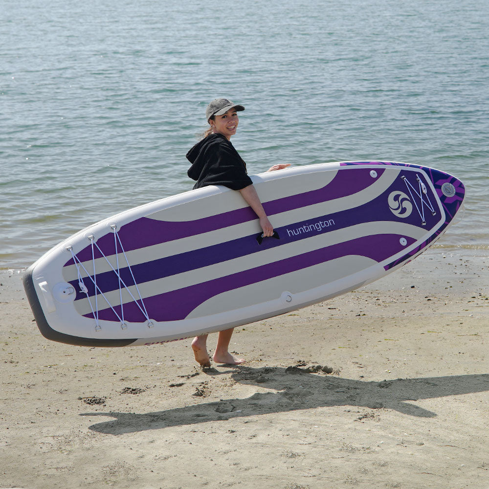 Person holding a purple and white Hurrican paddle board on a beach.