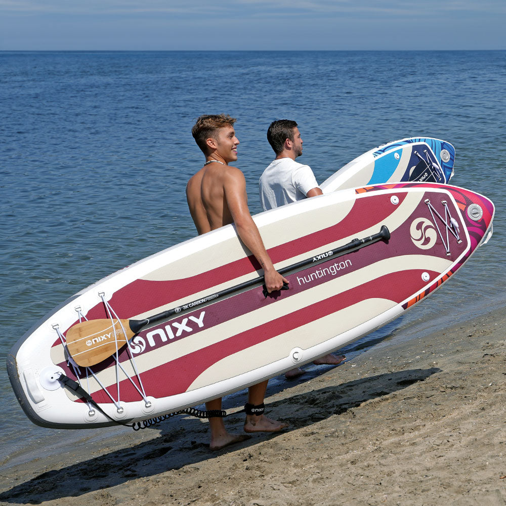 Two people carrying a red and white NIXY paddleboard on a beach.