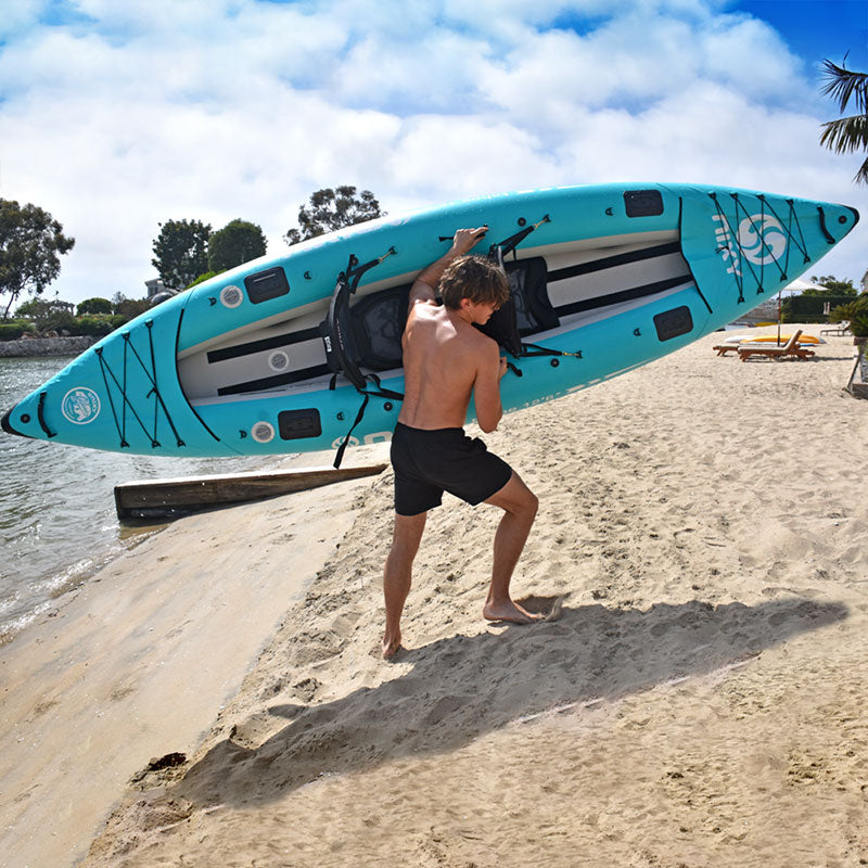 Man carrying a blue paddleboard on a sandy beach with trees and water in the background