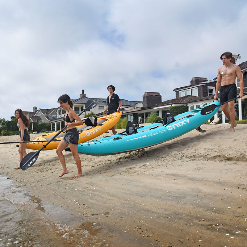 People carrying inflatable kayaks on a sandy beach with houses in the background
