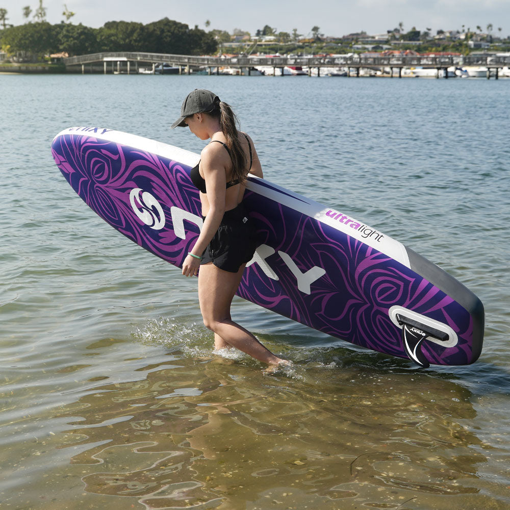 Woman carrying a purple and white inflatable paddle board with 'FRTY' branding through shallow water.