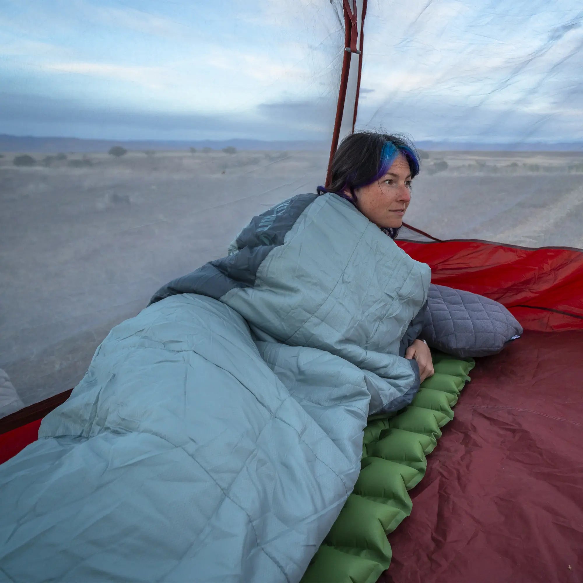 Person lying in a sleeping bag inside a hot air balloon with a scenic landscape below.