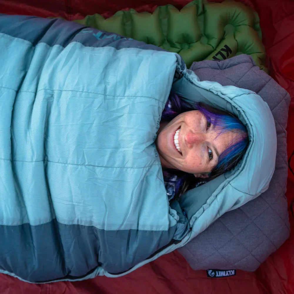 Person with colorful hair peeking out from a blue sleeping bag on a red sleeping pad.