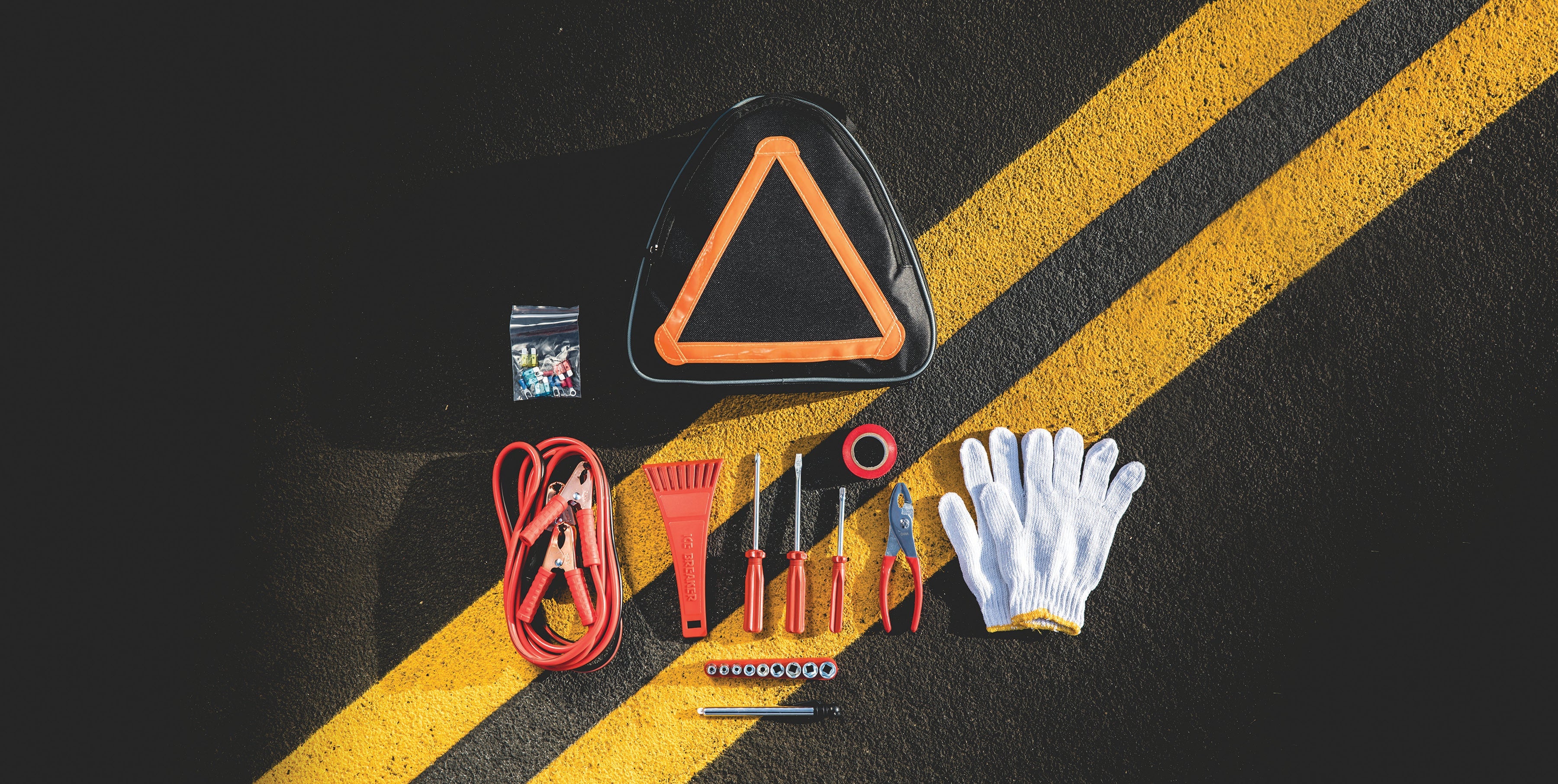 Collection of car emergency tools including a triangle warning sign, gloves, and jump leads on a black surface with yellow stripes.