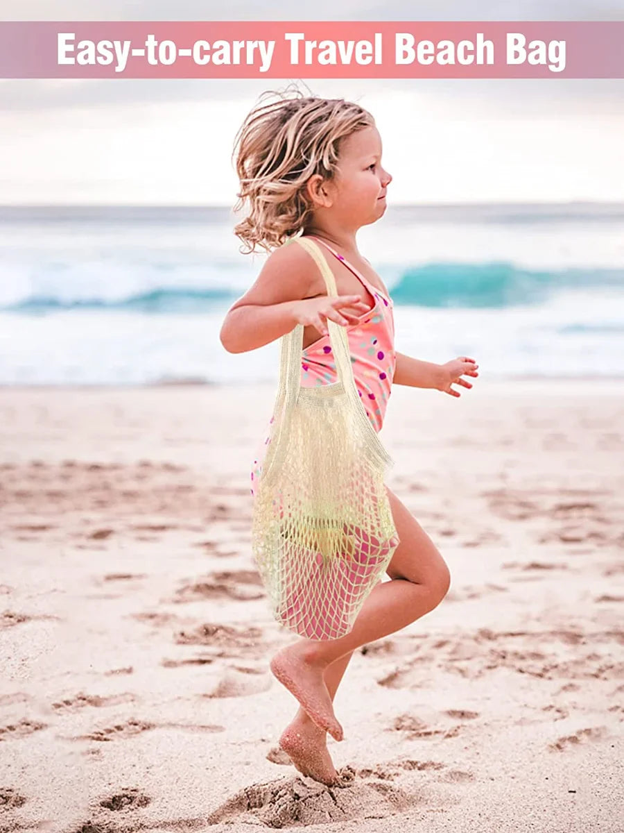 Child holding a yellow mesh beach bag on a sandy beach with ocean in the background.
