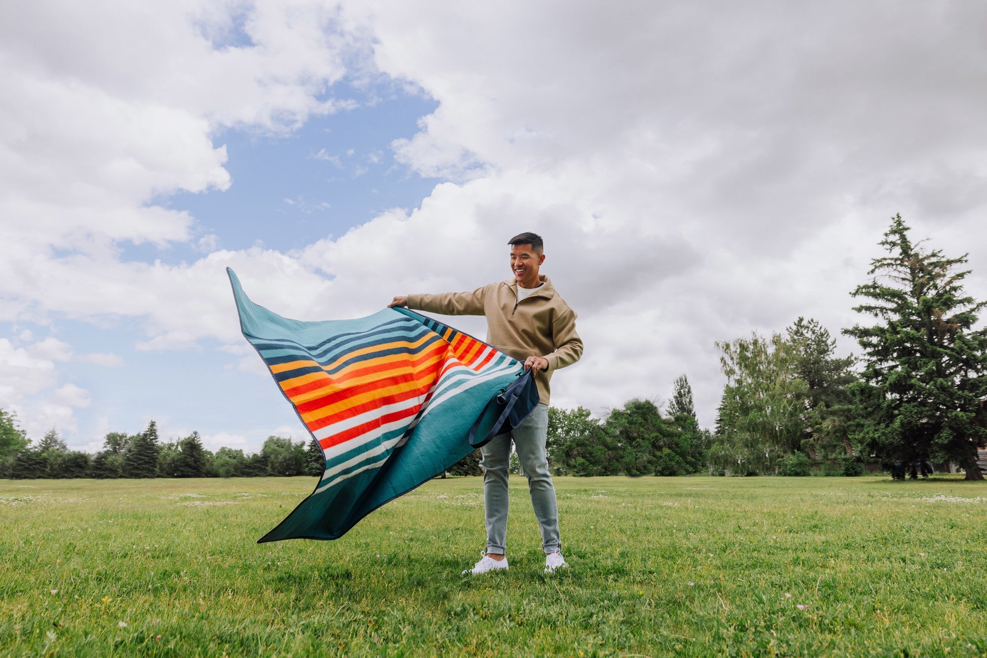 Person holding a colorful striped blanket in a grassy field with trees and blue sky.