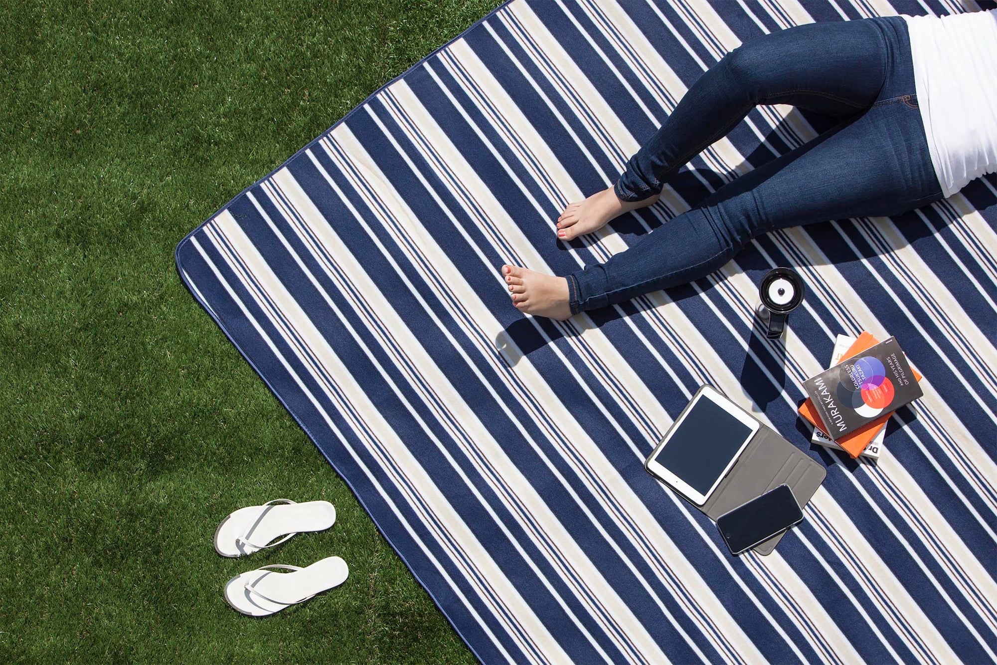 Person lying on a striped picnic blanket with electronic devices on grass