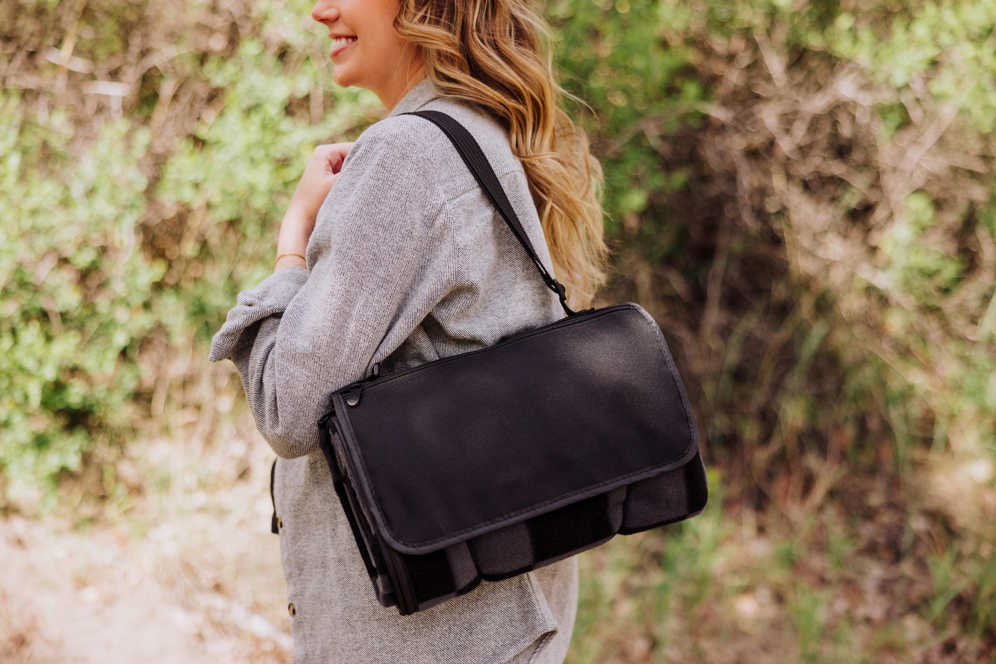 Woman carrying a black leather bag outdoors with greenery in the background