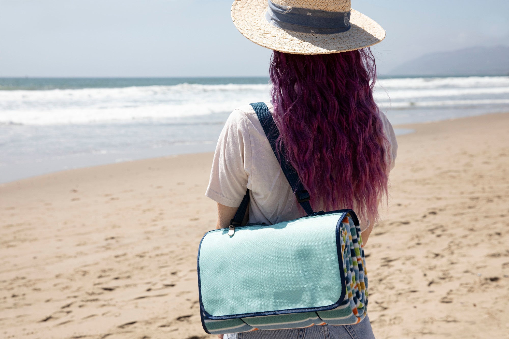Person with pink hair and a straw hat standing on a beach, holding a light blue bag.