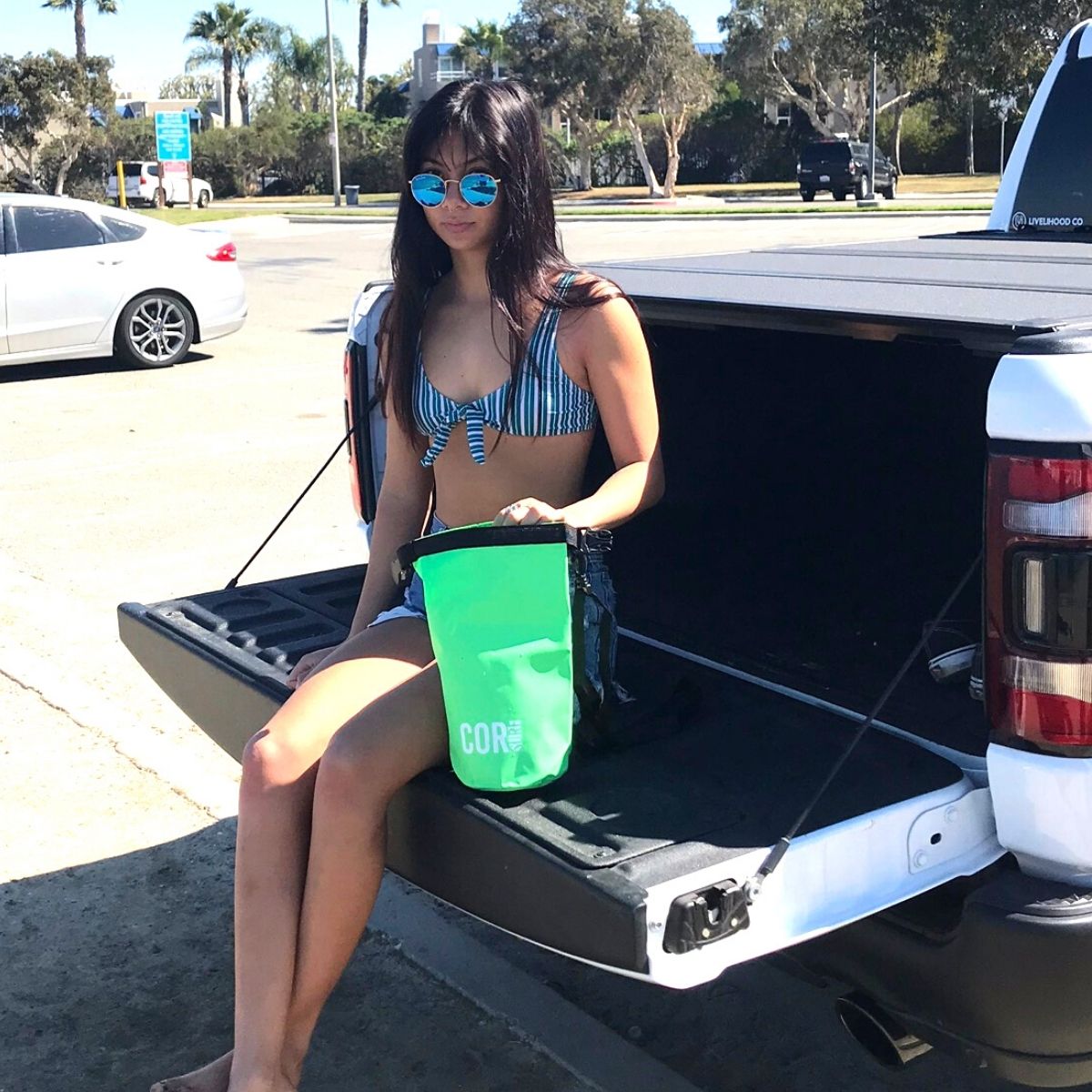 Woman sitting in a car trunk with a green bucket labeled 'CLORI' on a sunny day.