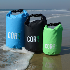 Three COR dry bags in blue, black, and green on a beach with ocean waves in the background.