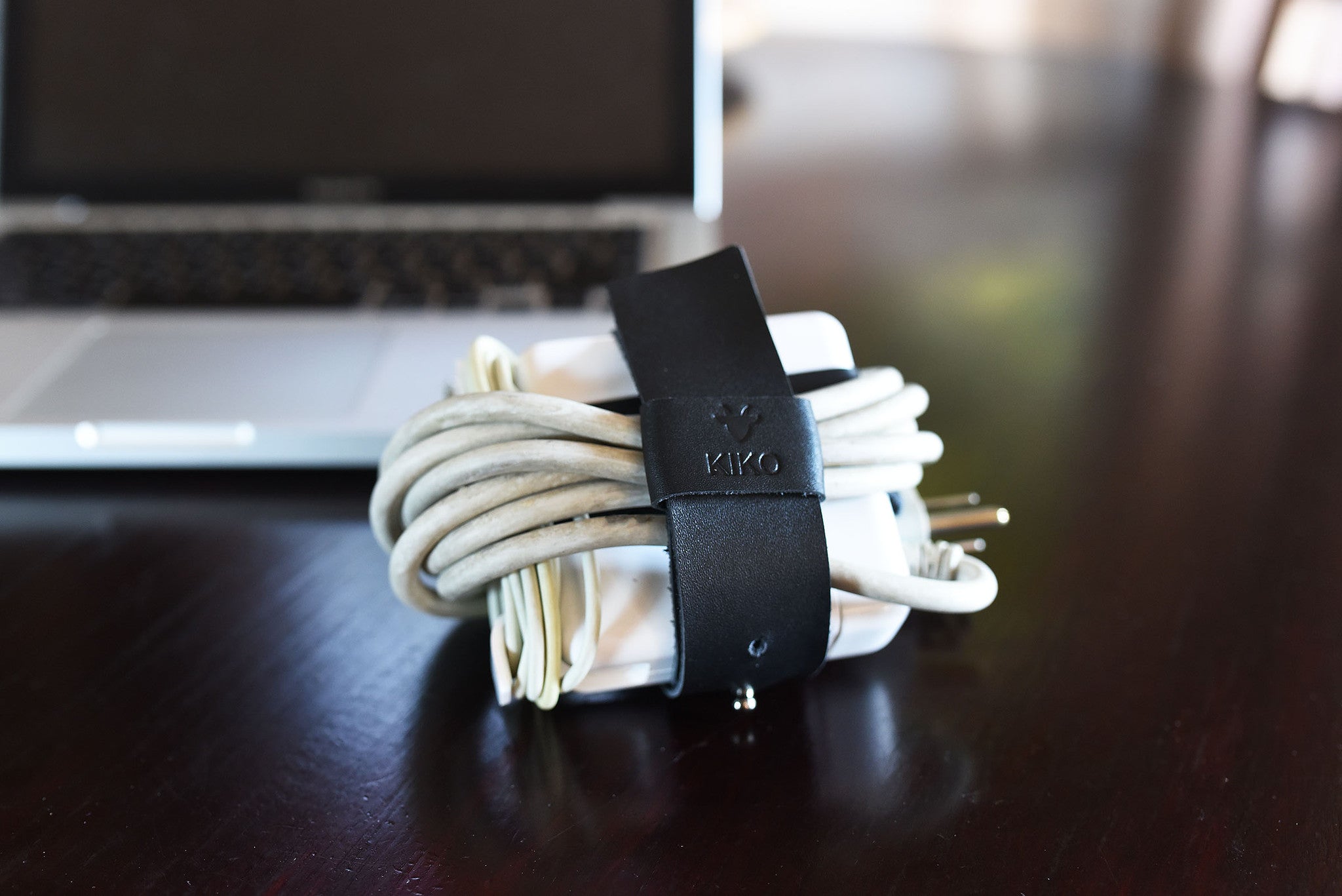 Cable organizer with white cables and black leather strap on a wooden desk with a laptop in the background.