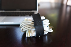 Cable organizer with white cables and black leather strap on a wooden desk with a laptop in the background.