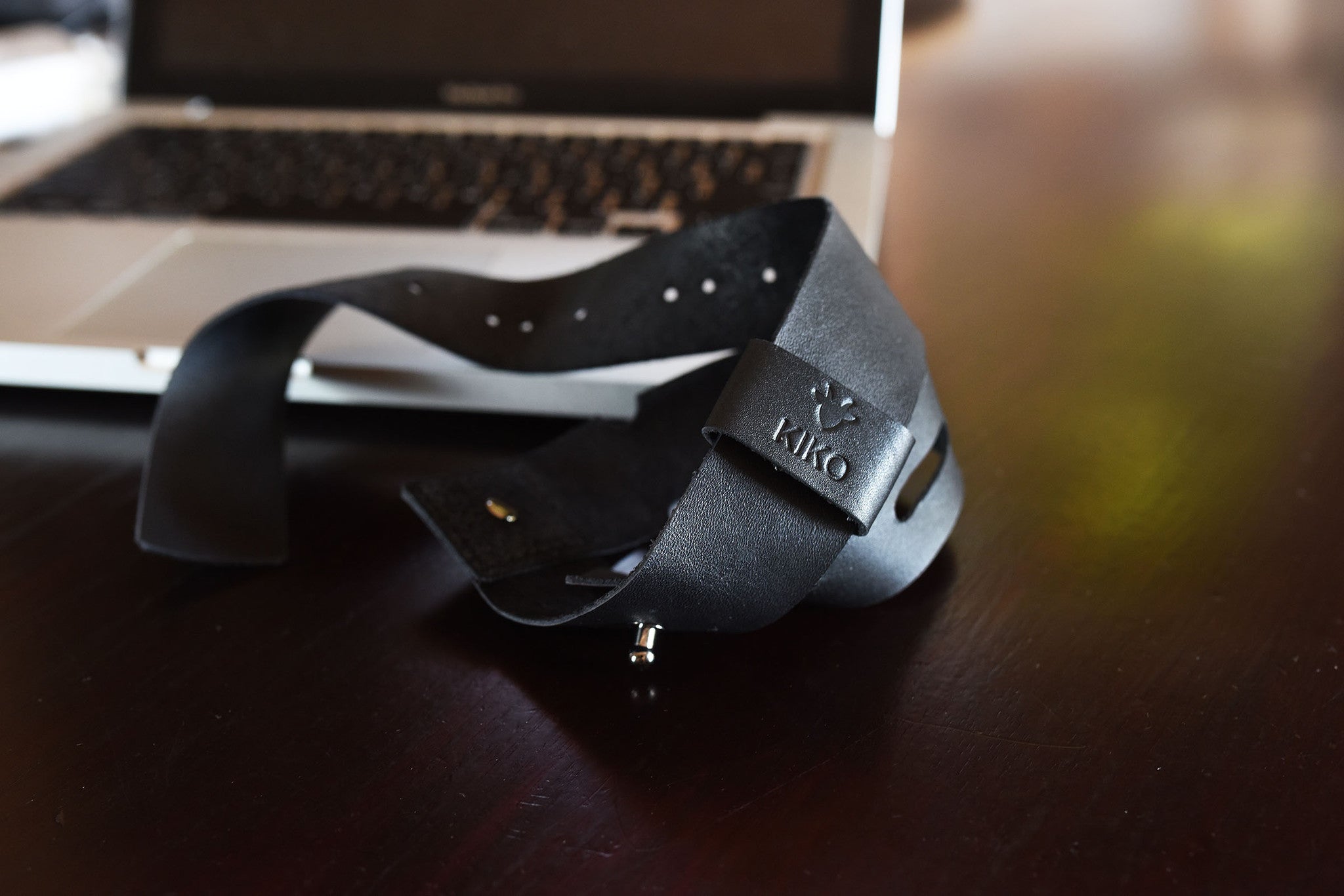 Black leather belt with a brand logo on a wooden surface next to a laptop.