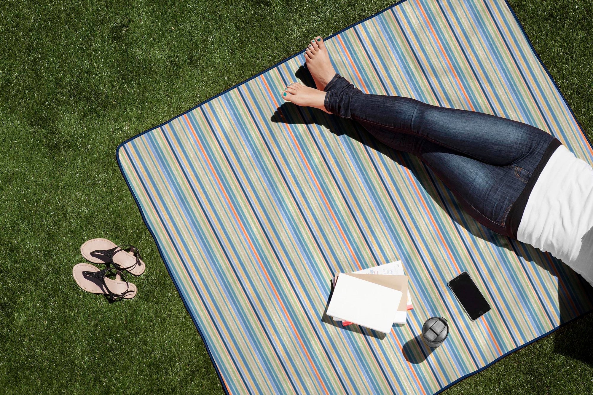 Person lying on a striped picnic blanket with a tablet and phone on a grassy area