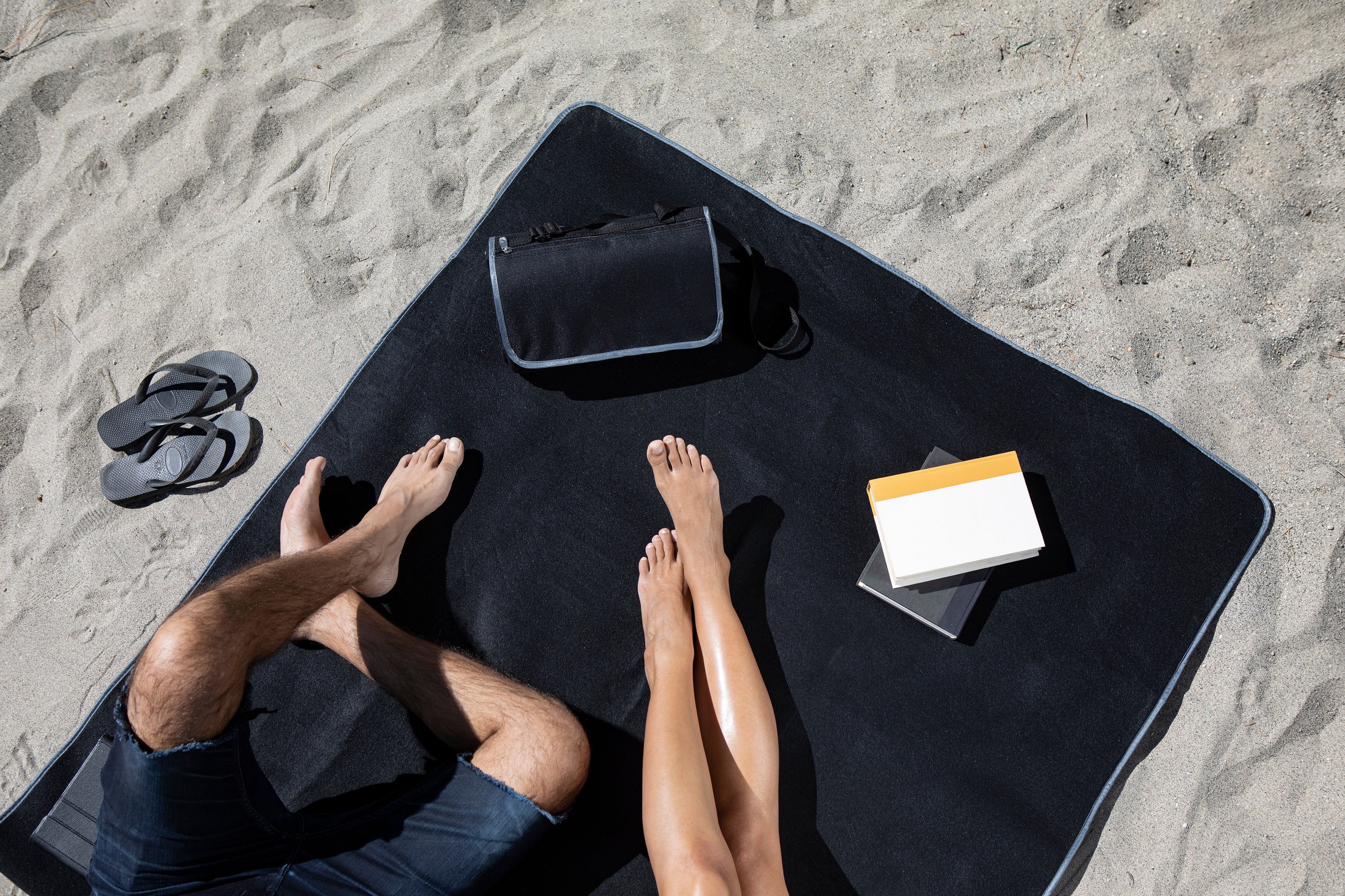 Person sitting on a black towel at the beach with a bag and books.