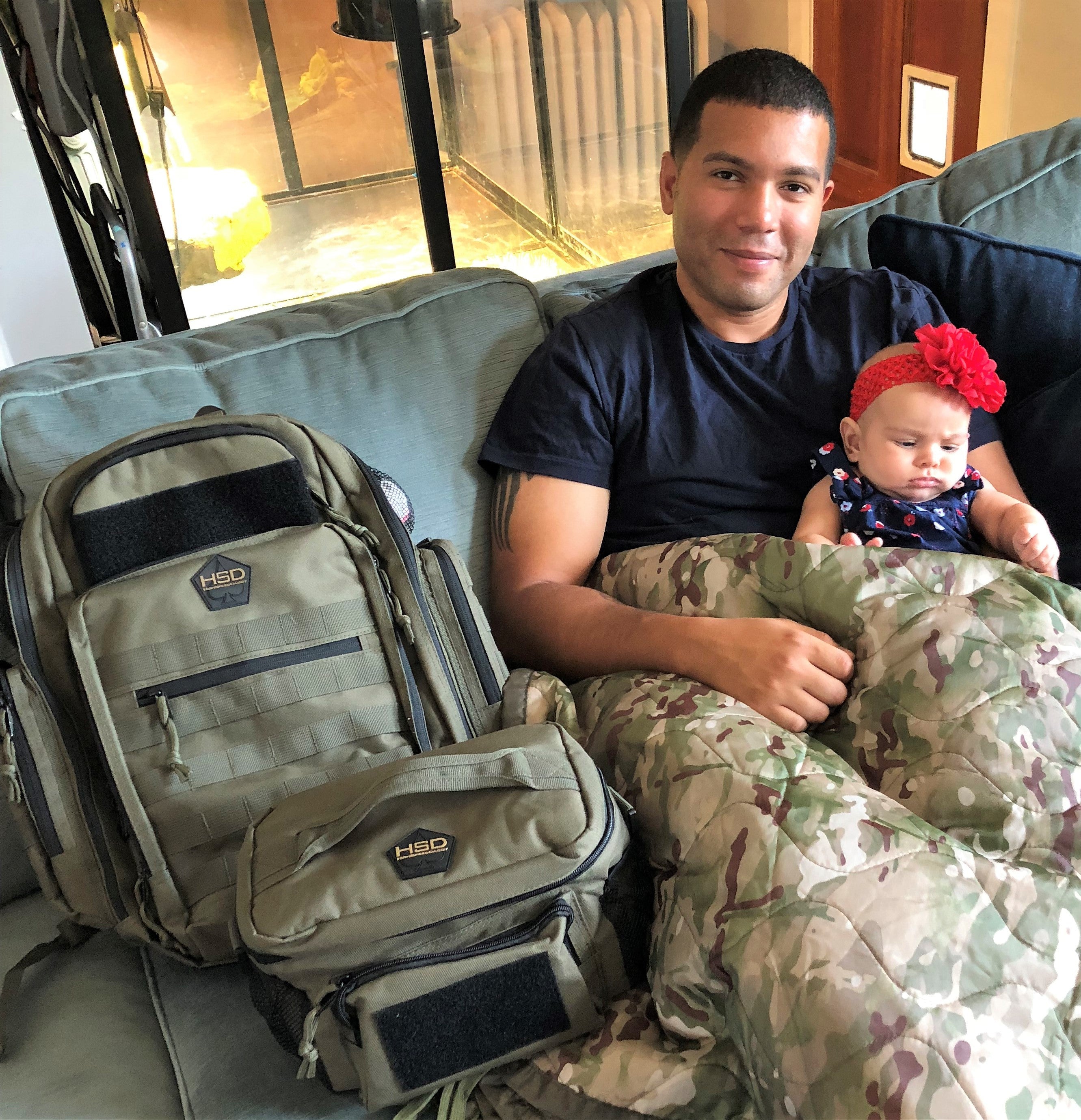 Man in camouflage pants holding a baby next to a large backpack on a couch.
