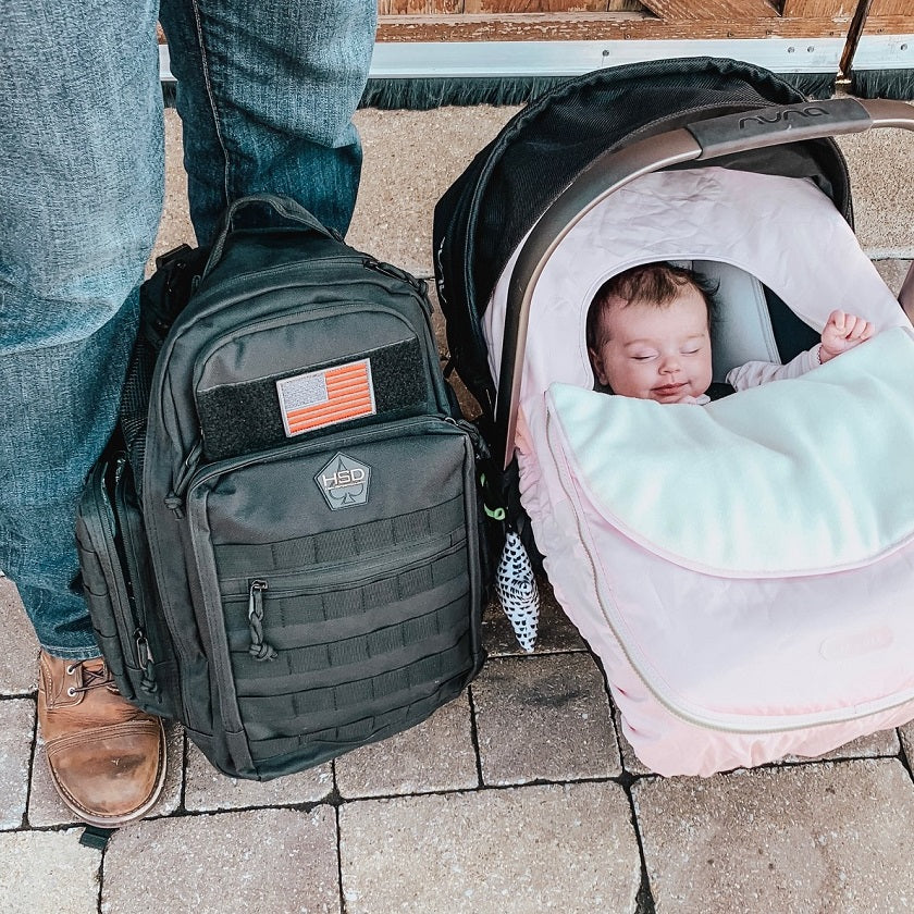 Person with a tactical backpack next to a baby in a stroller on a paved surface.