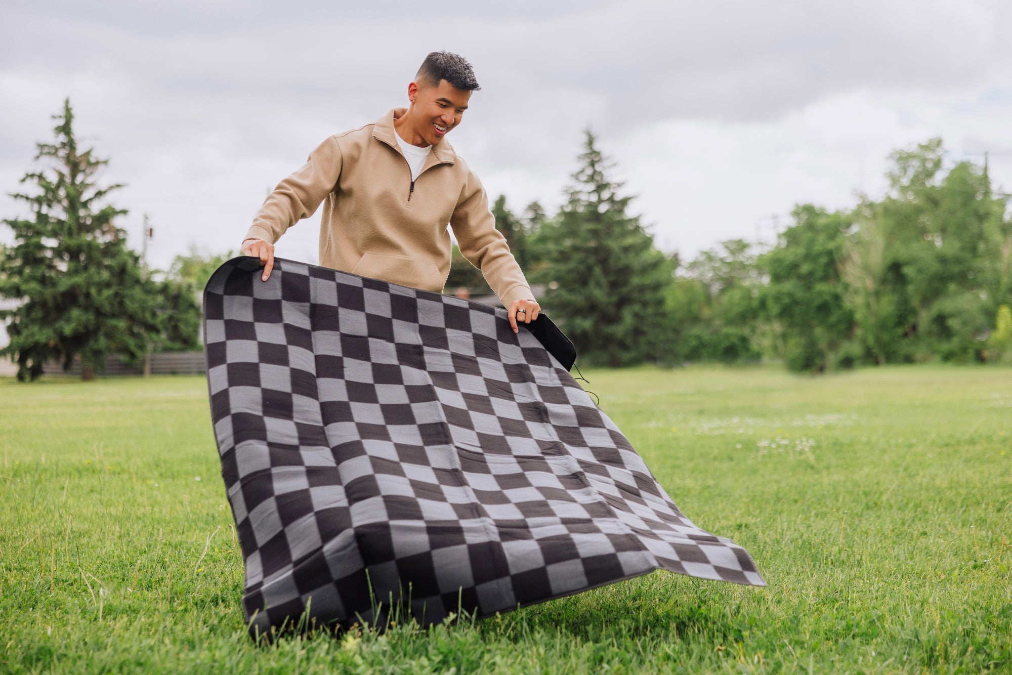 Person holding a black and gray checkered blanket outdoors in a grassy area with trees in the background.