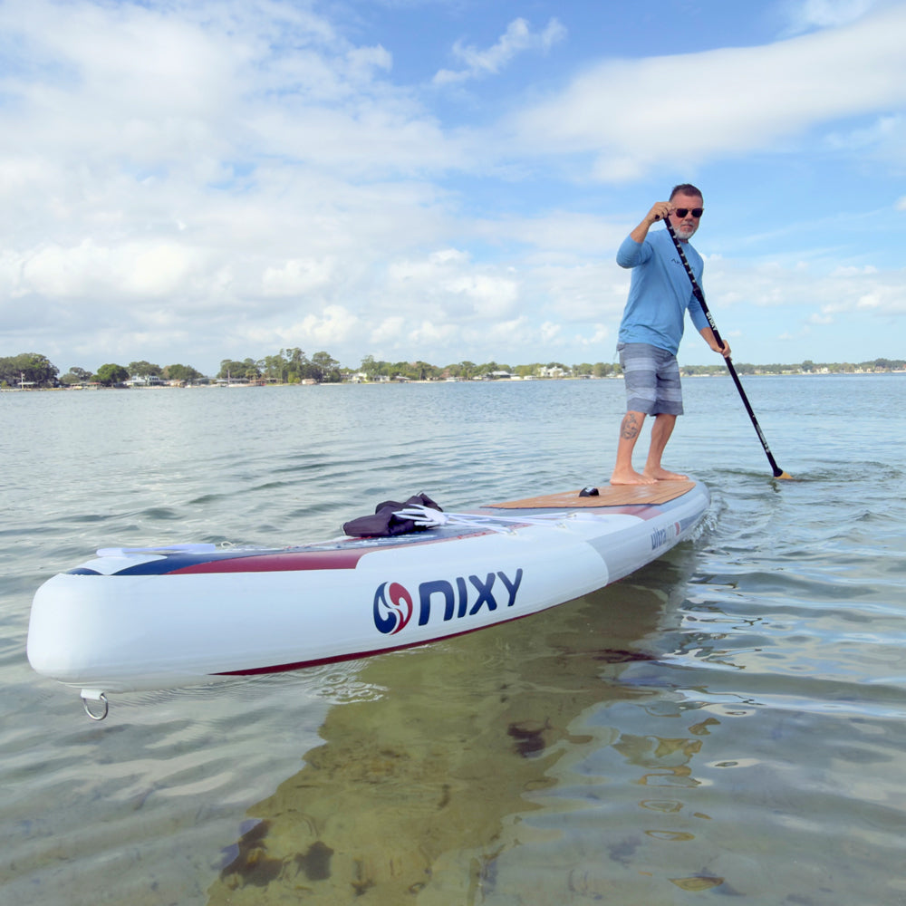 Person paddleboarding on a NIXY board in a calm body of water with trees and buildings in the background.