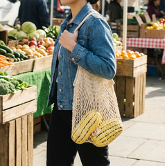 Person holding a string bag with vegetables at an outdoor farmers market.