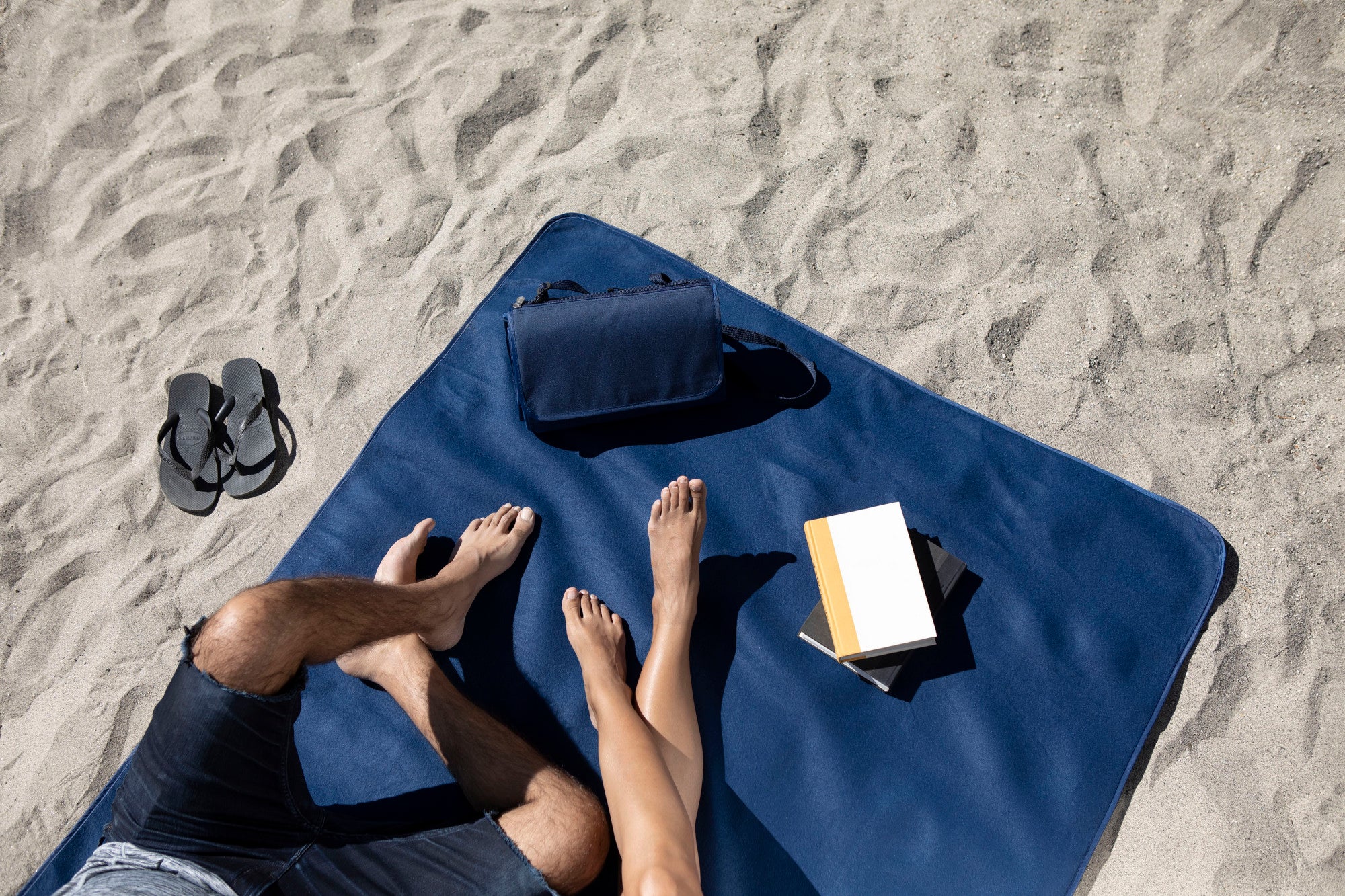 Two people lying on a blue towel with a bag and book on a sandy beach.