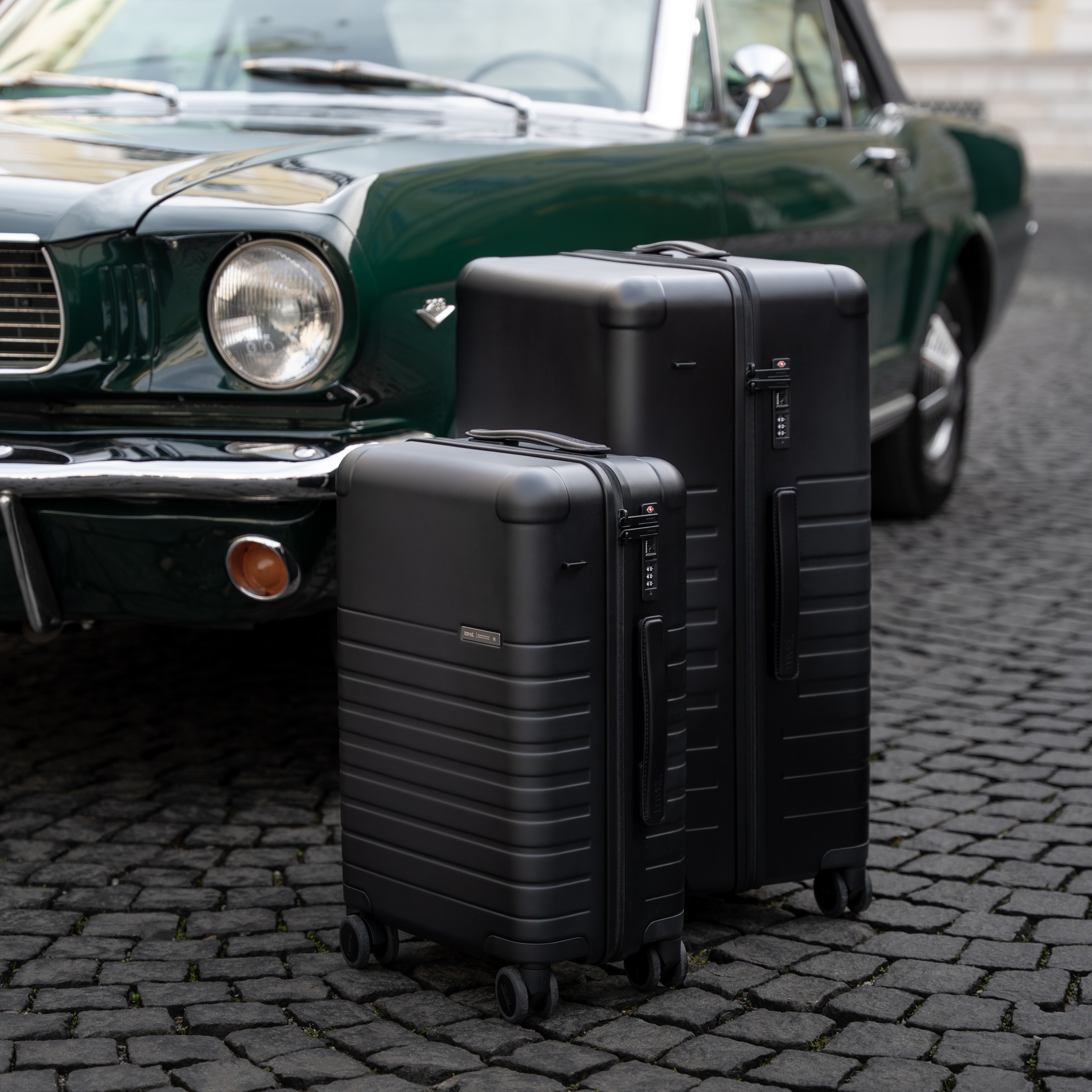 Two black suitcases on a cobblestone street with a classic green car in the background.