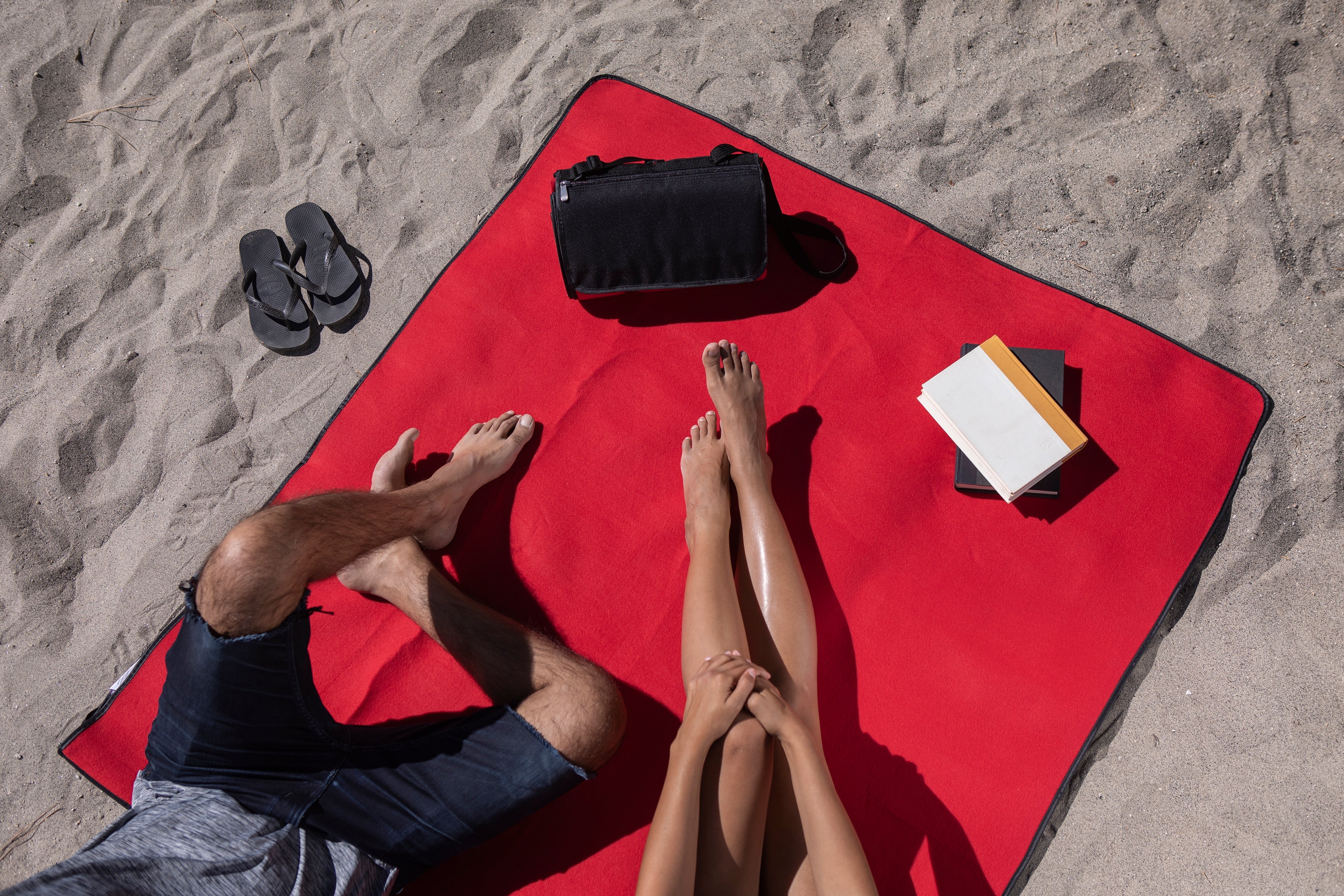Two people on a red towel at the beach with a bag and book.